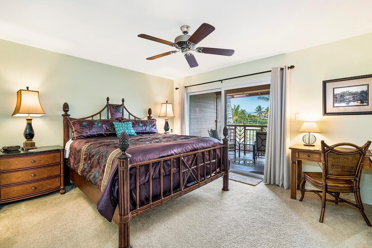 A bedroom with a bed, bedside lamps, a desk with a chair, a ceiling fan, and a view of the outdoors.