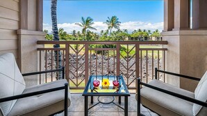 Ein Balkon mit zwei gepolsterten Stühlen, einem Glastisch mit zwei Getränken und einer Blume, und Blick auf Palmen sowie einen klaren Himmel.