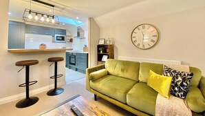 A modern living room with a green sofa, a large wall clock, and a kitchen in the background. - 2 bedroom accommodation in Cirencester (Cirencester)