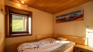 A bedroom with a wooden bed, a window with a view of greenery, a painting of a mountain landscape, and a nightstand with a lamp. - Small apartment on the ski slopes (Gryon)