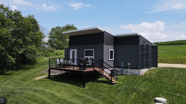 A modern two-story house with a deck, outdoor seating, and a staircase leading to the entrance. -  Quiet countryside shipping container home. Relax or enjoy outdoor recreation  (Westby)