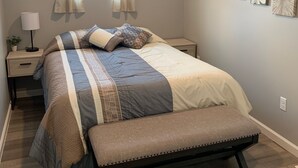 A bedroom with a bed, a bench, a nightstand, a lamp, and wall art. -  Quiet countryside shipping container home. Relax or enjoy outdoor recreation  (Westby)