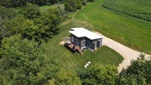 A modern house with a deck and outdoor seating, surrounded by lush greenery and fields. -  Quiet countryside shipping container home. Relax or enjoy outdoor recreation  (Westby)