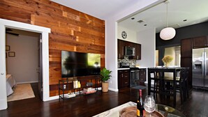 Un saló modern amb paret de fusta, televisió de pantalla plana, zona de menjador amb taula i caires, i cuina amb armaris foscos i electrodomèstics d'acer inoxidable.