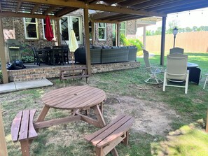 Eine überdachte Terrasse mit Holztisch und Bänken, einem Grill, Sitzgelegenheiten im Freien und Blick auf den Garten.