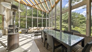Un amplio salón con ventanales, una gran mesa de vidrio con sillas, un sofá y una zona de barbacoa.