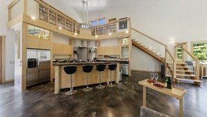 Una cocina moderna con una isla central, taburetes de bar, una mesa de comedor con una botella y copas, y una escalera que sube a un nivel superior.