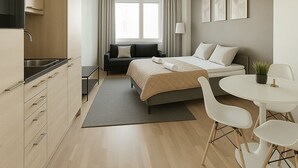 A modern hotel room with a kitchenette, a bed with a beige duvet, a bedside table with a lamp, a round dining table with chairs, and a window with curtains. - City Home Finland Arena Studio (Tampere)