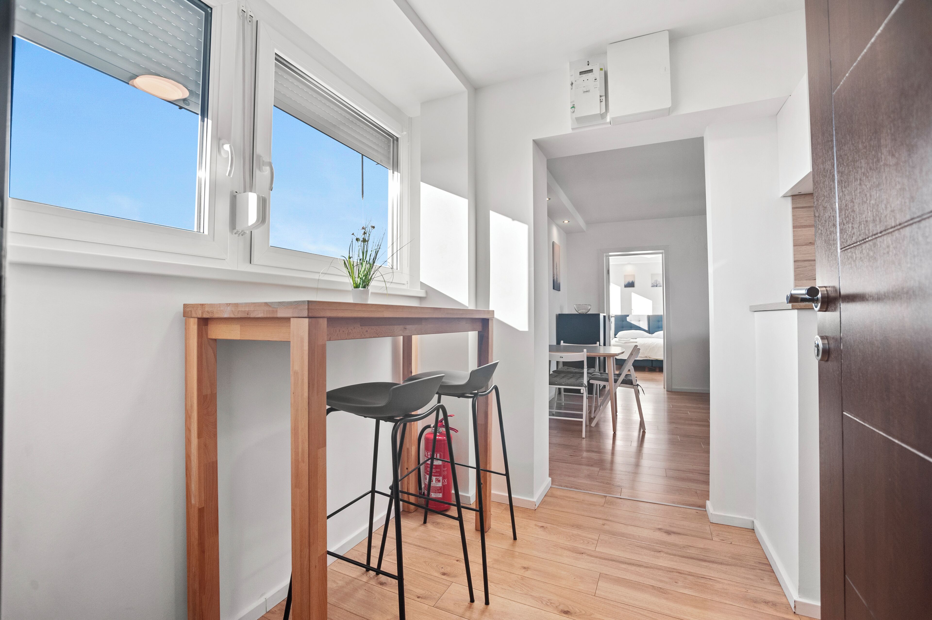 A modern kitchen with a wooden table and two bar stools, a window with blinds, and a visible fire extinguisher.