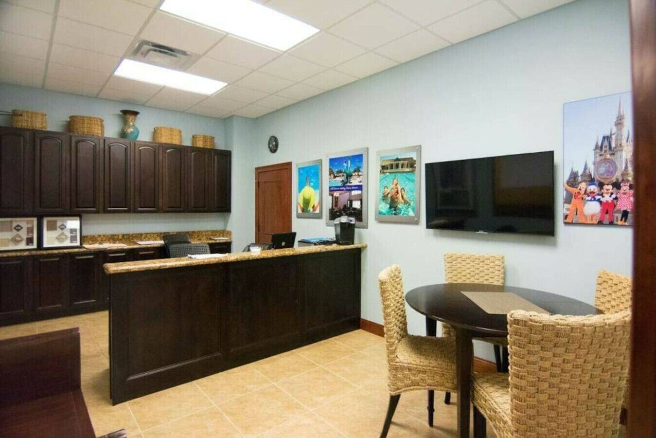 Uma cozinha com armários de madeira escura, bancada de granito, mesa de jantar com cadeiras de vime e uma TV de tela plana fixada na parede.