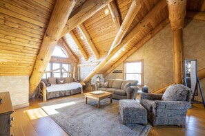 Een ruime loft met hoge plafonds, houten balken, een groot raam en een gezellige zithoek met een bank en fauteuils.