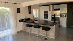 Cocina moderna con una isla central, taburetes de bar, electrodomésticos de acero inoxidable y un amplio ventanal con cortinas translúcidas.