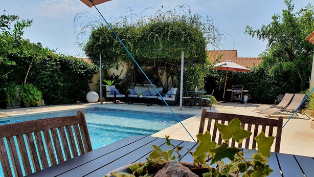 Un área junto a la piscina con mesa de madera, planta en maceta, sillas de descanso y una parrilla.