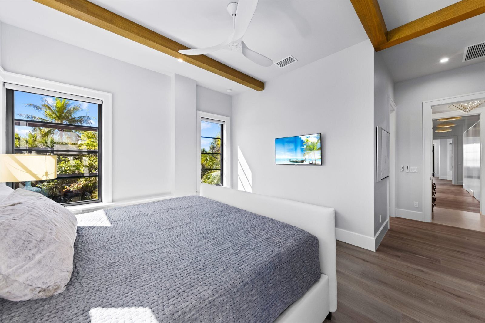 A modern bedroom with a large bed, a ceiling fan, and a view of palm trees through a window.