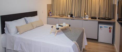 A compact hotel room with a bed, bedside table, and a kitchenette area.