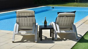 Två vita loungestolar placerade mittemot en swimmingpool, med en flaska och ett glas på ett litet bord mellan dem.
