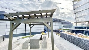 En utomhusplats med en pergola, sittbänkar och en modern byggnad i bakgrunden.