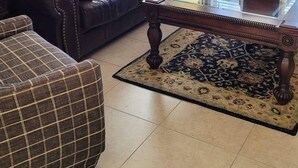 Ein Wohnzimmer mit einem dunkelbraunen Ledersofa, einem Glastisch, einem karierten Hocker und einem floralen Gemälde an der Wand.