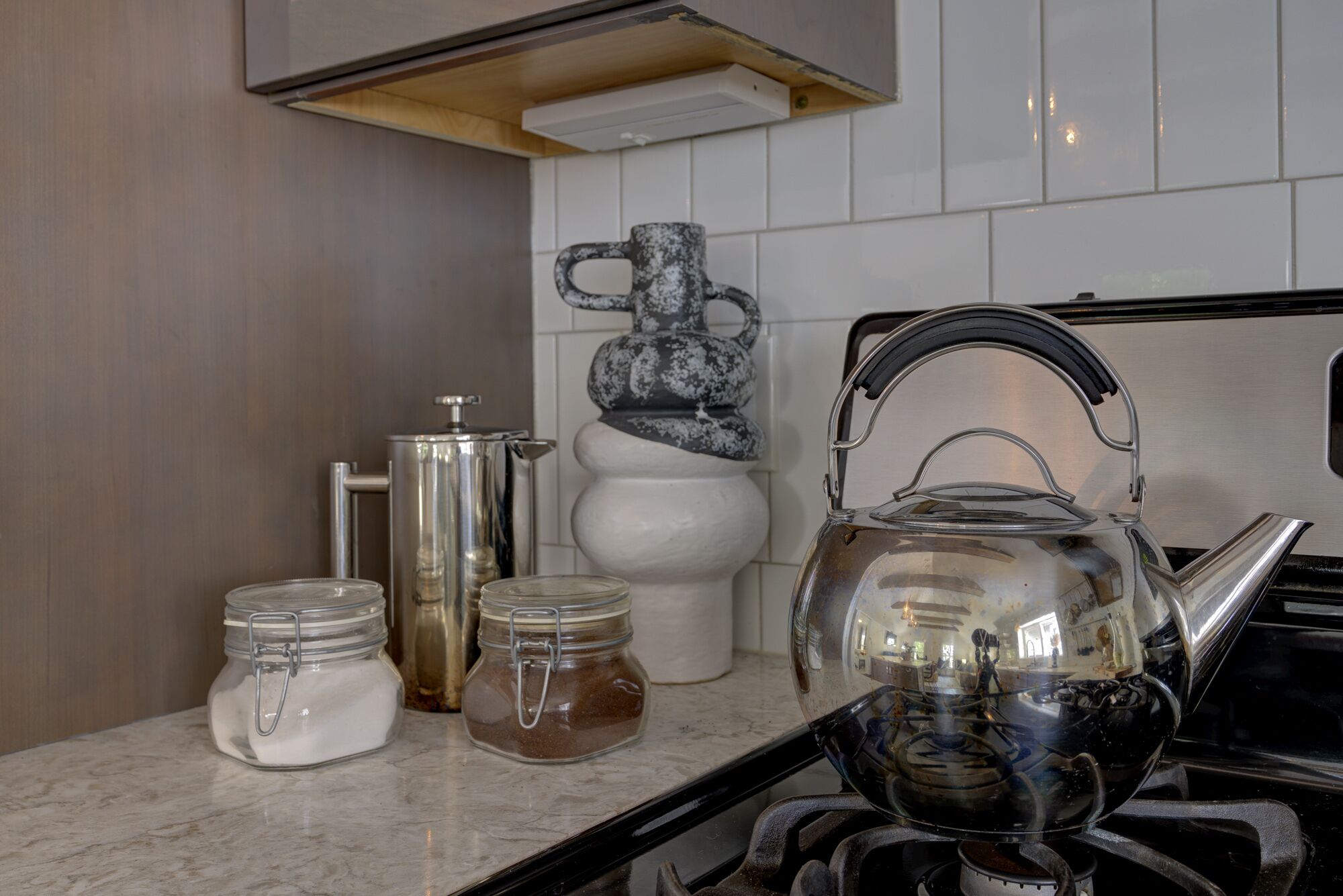 Une cuisine équipée d’une cuisinière à gaz, d’une bouilloire, de deux bocaux avec couvercles, d’une cafetière à piston et d’un vase décoratif.