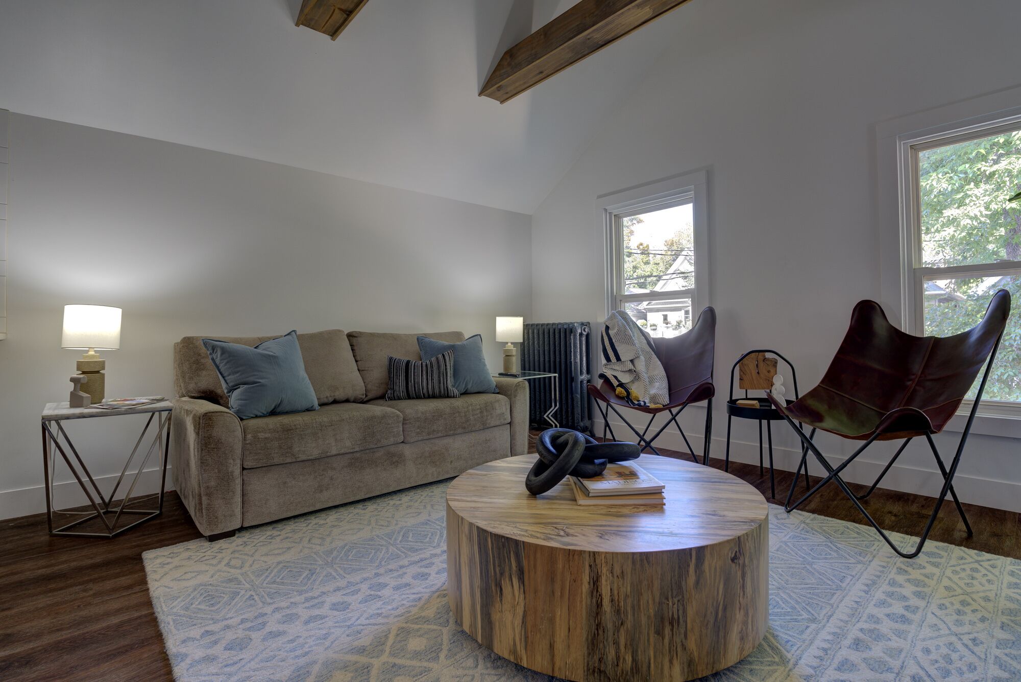 A living room with a sofa, a round coffee table, and a chair. There is a window with a view of trees and a radiator.