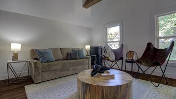 A living room with a sofa, a round coffee table, and a chair. There is a window with a view of trees and a radiator.