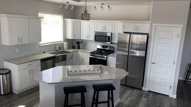 A modern kitchen with white cabinets, a stainless steel refrigerator, a microwave, and a gas stove. There is a large island with stools and a window providing natural light. - Brand new, 3 bedroom piece of  paradise! (Grand Junction)