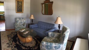 Ein Wohnzimmer mit einem blauen Sofa, gemusterten Sessel und einem Glastisch, sowie zwei Lampen.