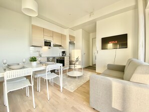 A modern kitchen with white cabinetry, a dining area with a white table and chairs, a grey sofa, and a flat-screen TV mounted on the wall. - City Home Finland Ratina Panorama (Tampere)