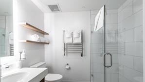 A modern bathroom with a white sink, toilet, and shower area. There are shelves with towels and a glass shower door.