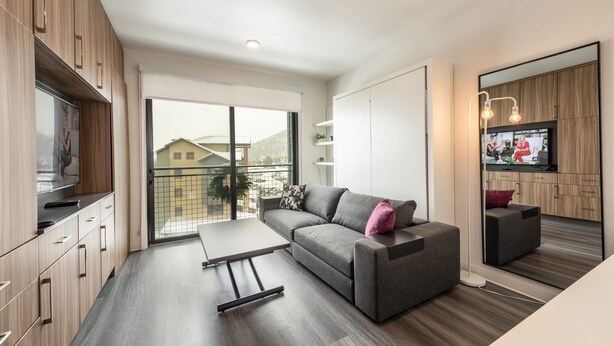 A modern living room with a sofa, a coffee table, and a TV. There is a balcony with a view of buildings and trees.