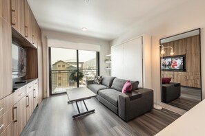 A modern living room with a sofa, a coffee table, and a TV. There is a balcony with a view of buildings and trees. - Studio Pad by Canyons Village Rentals (Park City)