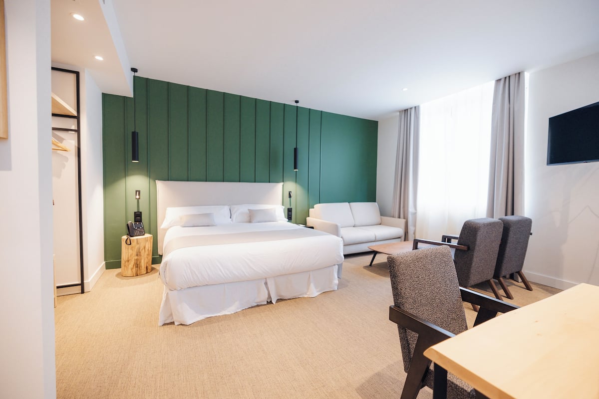 A modern hotel room with a large bed, a sofa, a dining table, and a TV mounted on the wall.