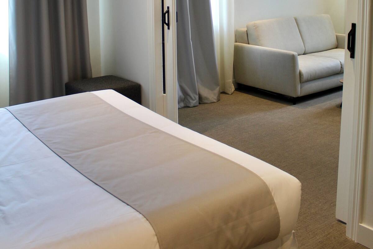 A hotel room with a large bed, a small seating area, and a door leading to another room.