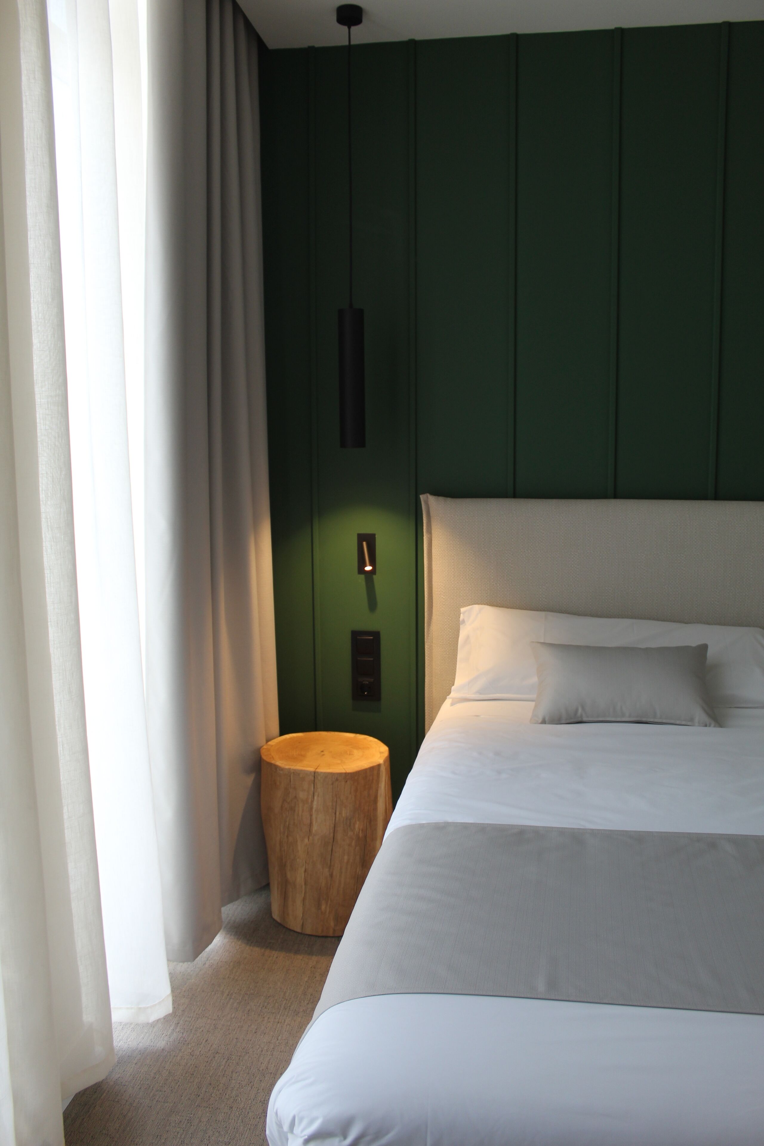 Una cama bien hecha con sábanas blancas, una mesita de madera redonda y una pared verde con un interruptor de luz.