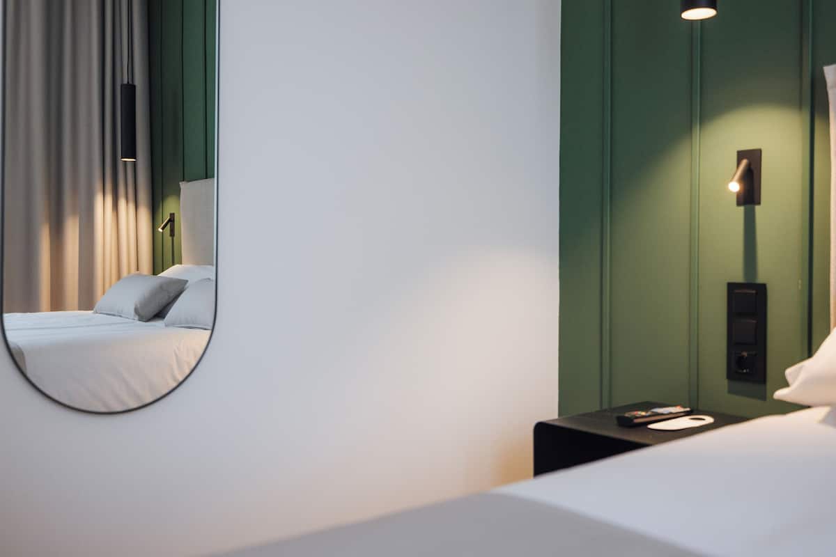 A hotel room with a bed, a nightstand, a mirror, and a wall-mounted light fixture.