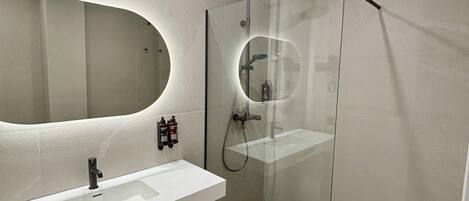 Un baño moderno con un espejo grande, un lavabo blanco y una zona de ducha con mampara de cristal.