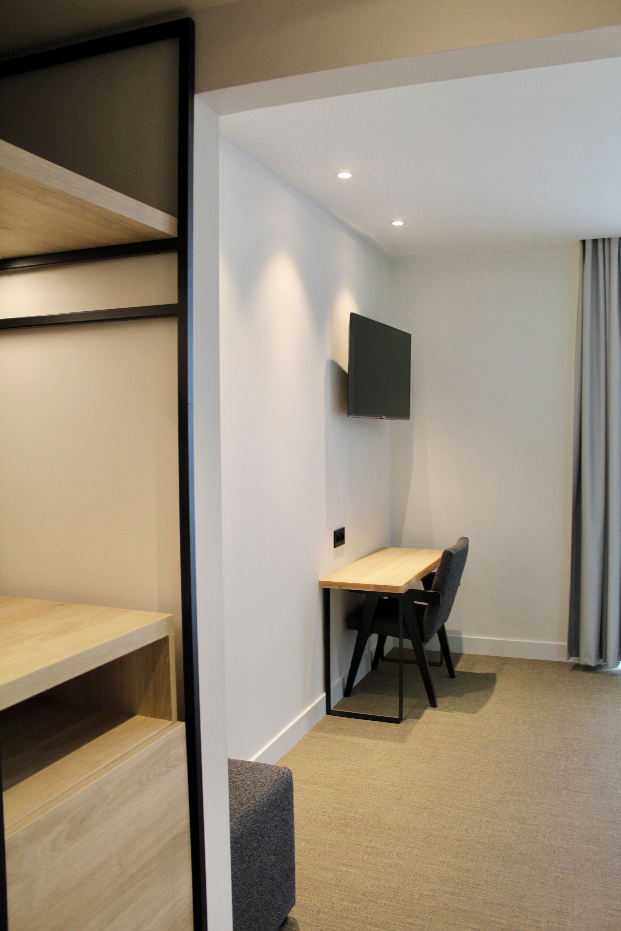 A modern hotel room with a wooden desk, a flat-screen TV mounted on the wall, and a comfortable chair.