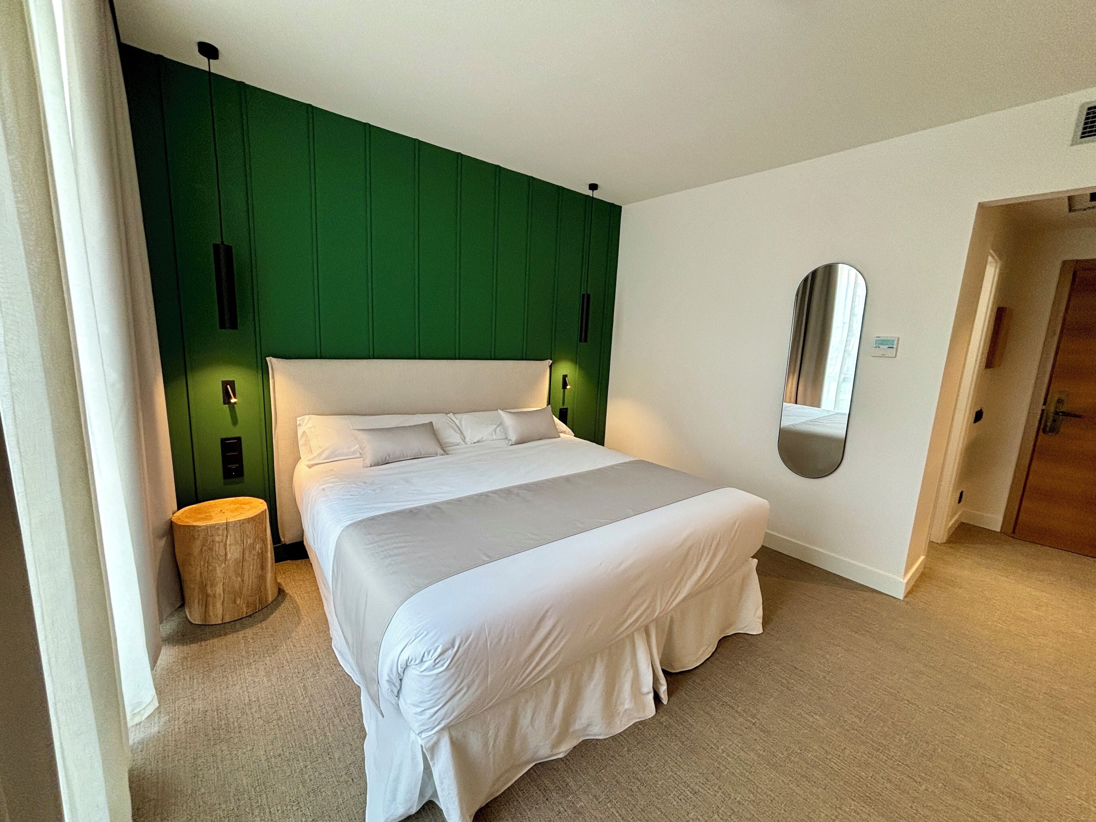 A neatly made bed with white linens in a room with green paneling on the walls and a large mirror on the wall.
