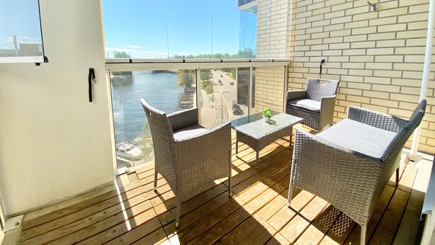Ein Balkon mit Korbmöbeln, einem Glastisch und Blick auf einen Fluss und Gebäude.