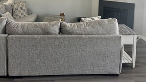 Un salon moderne avec un canapé d’angle gris, un ventilateur de plafond, un téléviseur à écran plat et une œuvre d’art encadrée représentant des planches de surf.
