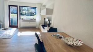A modern living room with a wooden dining table, a sofa, and a large window providing a view of the outdoors. - Sunside vacation apartment in the heart of Winterberg (Winterberg)