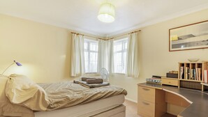 Un dormitorio con cama, mesita de noche, lámpara, ventana con cortinas y un escritorio con silla.