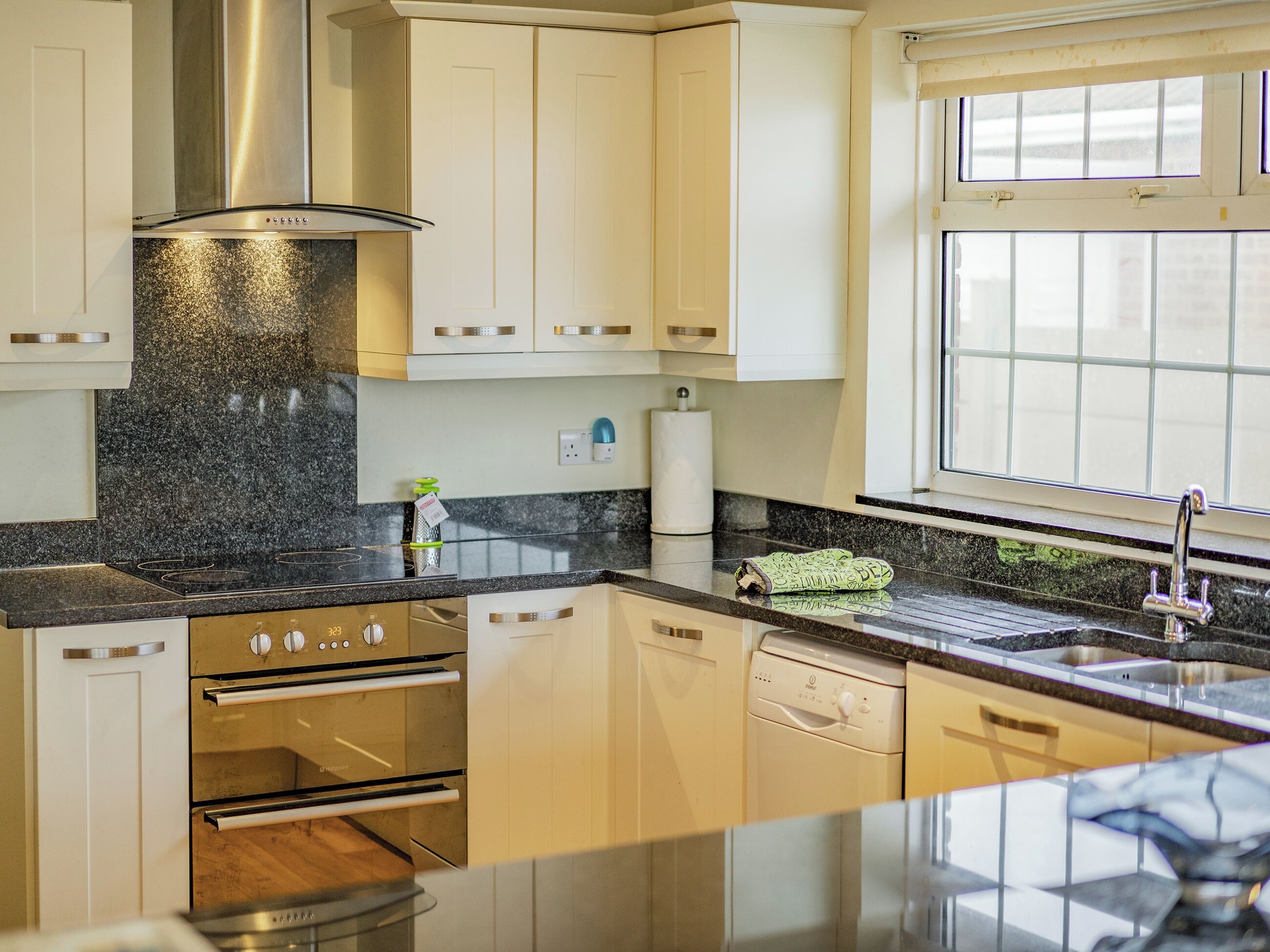 Una cocina moderna con gabinetes blancos, una campana extractora de acero inoxidable, horno empotrado, lavavajillas y ventana que proporciona luz natural.