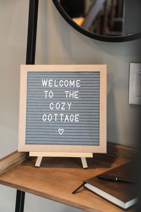 A framed welcome sign with the text "WELCOME TO THE COZY COTTAGE" on a wooden table.