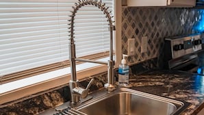 A kitchen with a stainless steel sink, a bottle of hand soap, and a window with blinds.