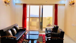 A living room with a sofa, chairs, a table, and a chandelier. There are windows with red curtains and a view outside. - High-rise, Most Private, No Noise, Room & Common Areas (Ghaziabad)