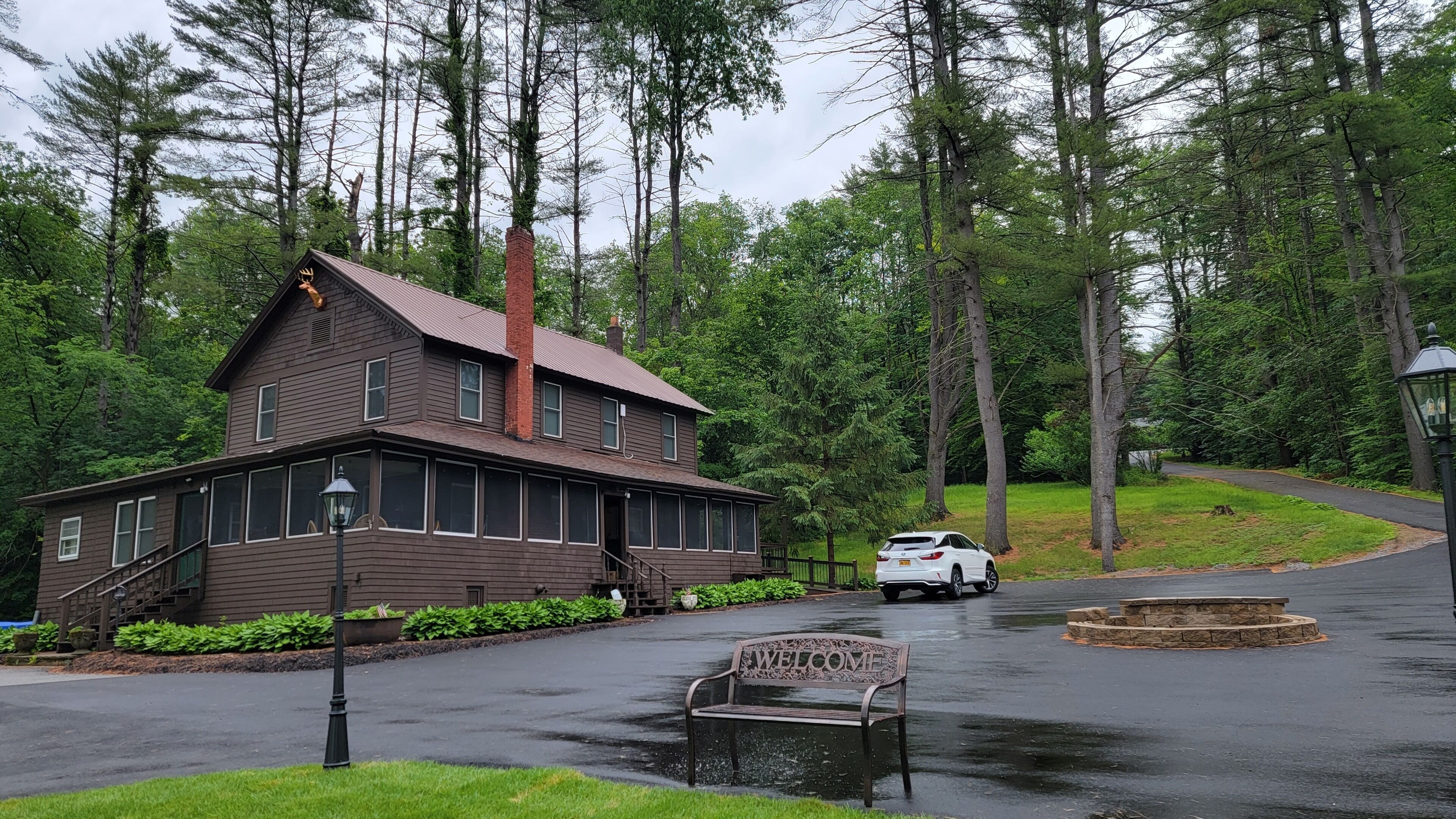 A two-story wooden building with a chimney, a bench with 'WELCOME' engraved, and a car parked in front.