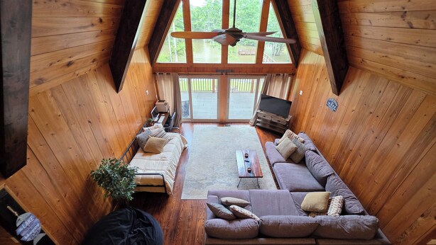 A cozy loft-style room with a wooden interior, a large window overlooking a deck, a ceiling fan, and a television on a wooden stand.