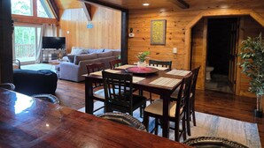 A wooden cabin interior with a dining table, chairs, a sofa, and a television.