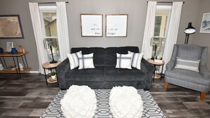 Ein Wohnzimmer mit einem dunkelgrauen Sofa, einem gemusterten Teppich und zwei gerahmten Motivationssprüchen an der Wand.